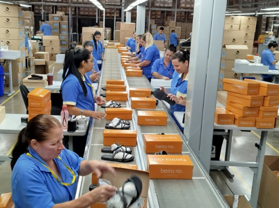 Equipe de produção em fábrica de calçados embalando pares em caixas laranja ao longo de uma linha de expedição